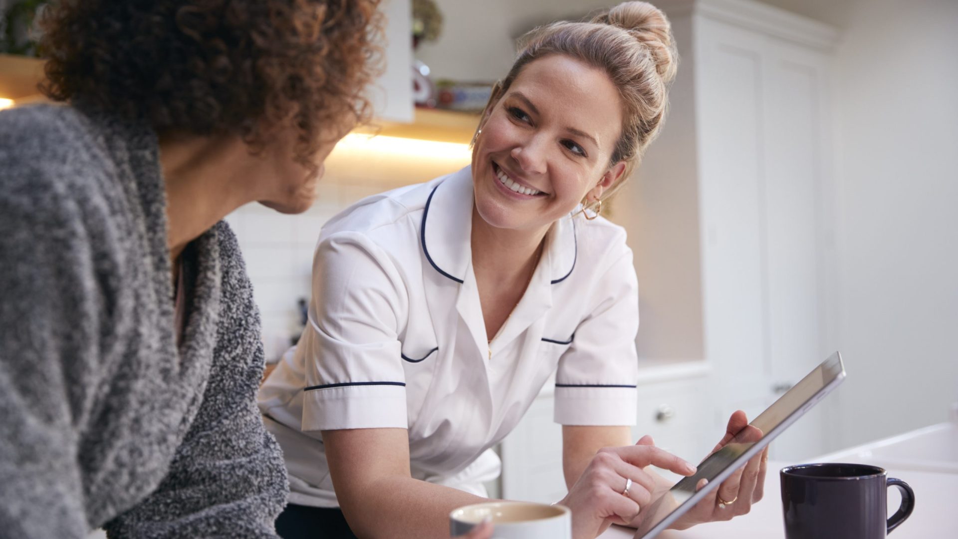 Mature Woman In Dressing Gown At Home Talking With Female Nurse With Digital Tablet