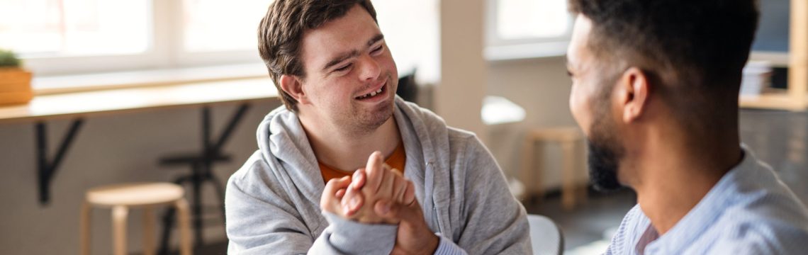 A young happy man with Down syndrome with his mentoring friend celebrating success indoors at school.