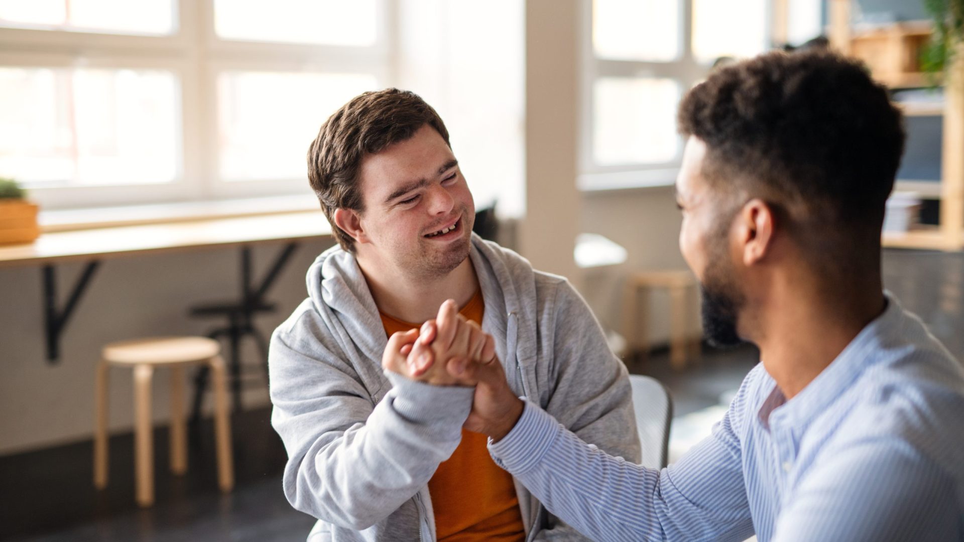 A young happy man with Down syndrome with his mentoring friend celebrating success indoors at school.
